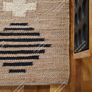 May include: A rectangular woven rug made of natural tan fibres featuring a large black circular pattern composed of horizontal lines and a white cross-shaped design at the top. The rug has a thick, textured weave and a visible border edge.