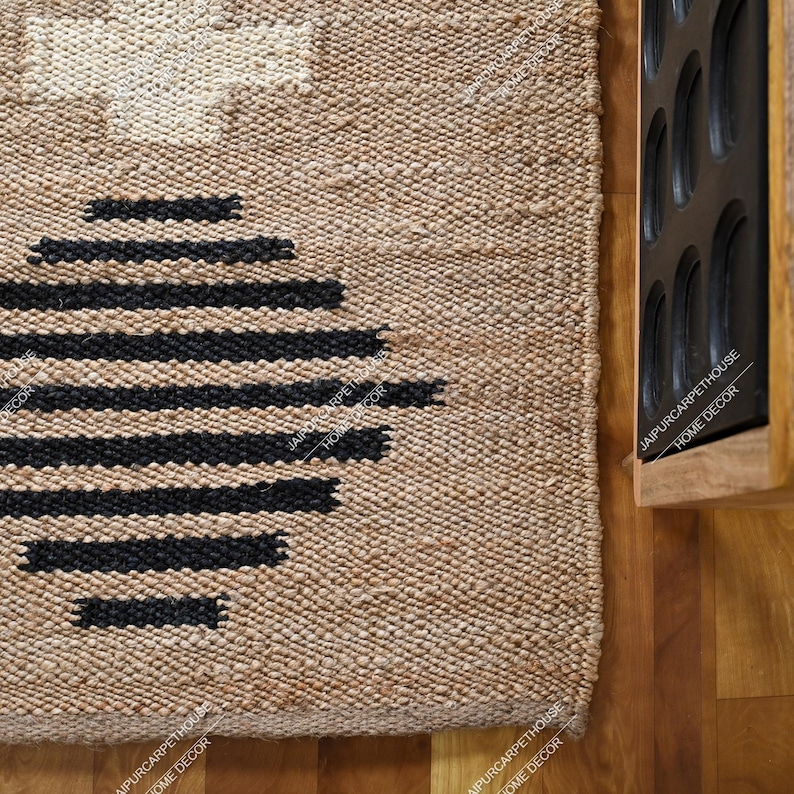 May include: A rectangular woven rug made of natural tan fibres featuring a large black circular pattern composed of horizontal lines and a white cross-shaped design at the top. The rug has a thick, textured weave and a visible border edge.