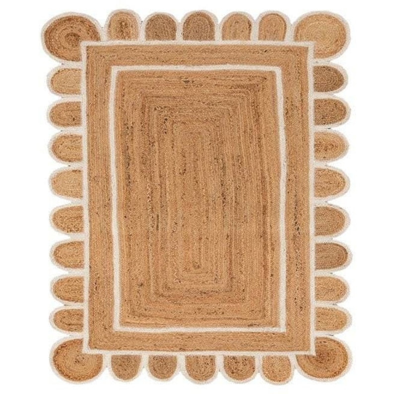 Puede incluir: Una alfombra de fibra natural con un borde blanco y un borde festoneado. La alfombra est&aacute; hecha de yute y tiene un color natural y terroso.