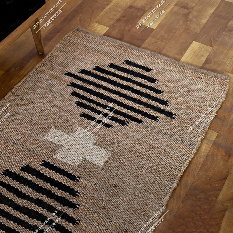 May include: A rectangular woven rug made of natural tan fibres featuring a central white cross shape flanked by two large black geometric patterns composed of horizontal parallel lines. The rug has a textured surface and a visible braided weave.