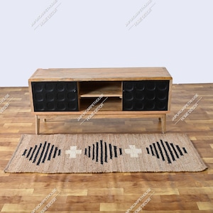 May include: A rectangular wooden media console with a natural light brown finish, featuring two black cabinet doors with a circular embossed pattern and an open centre shelf. Below sits a rectangular jute rug with a black geometric diamond pattern.