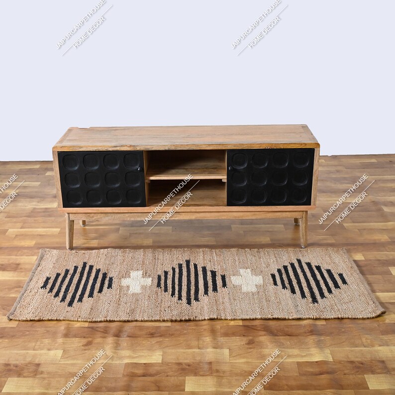 May include: A rectangular wooden media console with a natural light brown finish, featuring two black cabinet doors with a circular embossed pattern and an open centre shelf. Below sits a rectangular jute rug with a black geometric diamond pattern.