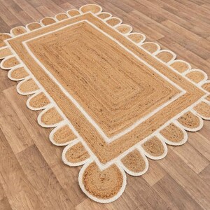 Puede incluir: Una alfombra de fibra natural con un borde blanco y un borde festoneado. La alfombra est&aacute; hecha de yute y tiene un color natural y terroso.
