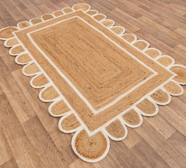 Puede incluir: Una alfombra de fibra natural con un borde blanco y un borde festoneado. La alfombra est&aacute; hecha de yute y tiene un color natural y terroso.