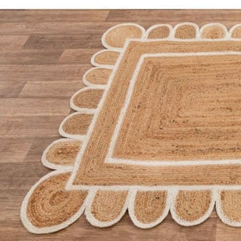 Puede incluir: Una alfombra de fibra natural con forma cuadrada y un borde blanco. La alfombra tiene un borde festoneado con un ribete blanco.