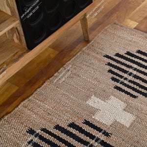 May include: A rectangular woven rug made of natural tan fibres featuring a repeating pattern of black horizontal lines and a central white cross shape. The rug is positioned on a wooden floor next to a wooden cabinet with black circular cutouts.