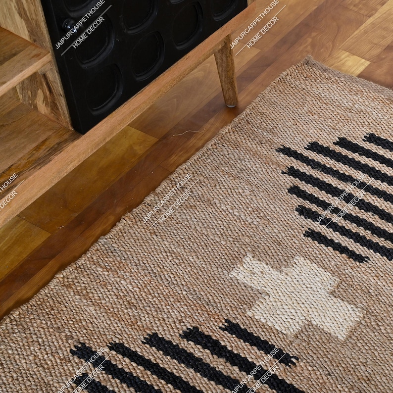 May include: A rectangular woven rug made of natural tan fibres featuring a repeating pattern of black horizontal lines and a central white cross shape. The rug is positioned on a wooden floor next to a wooden cabinet with black circular cutouts.