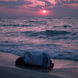 May include: A person lies face down on a sandy beach with their arms wrapped around their body. The ocean waves lap at the shore in the background. The sky is a vibrant pink and orange color as the sun sets in the distance.
