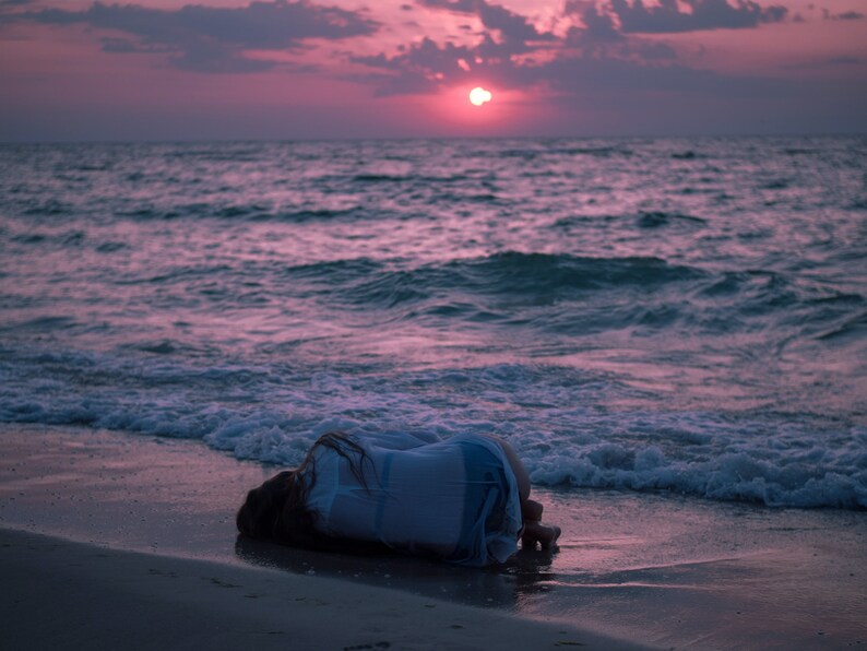 May include: A person lies face down on a sandy beach with their arms wrapped around their body. The ocean waves lap at the shore in the background. The sky is a vibrant pink and orange color as the sun sets in the distance.
