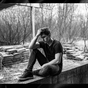May include: A young person sits on a concrete ledge with their head in their hands. They are wearing a black t-shirt and jeans. The background is a blurry image of trees and a pile of wood.