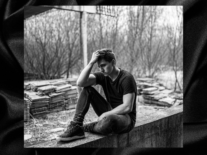 May include: A young person sits on a concrete ledge with their head in their hands. They are wearing a black t-shirt and jeans. The background is a blurry image of trees and a pile of wood.