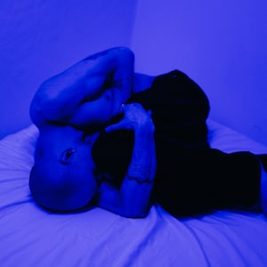 May include: Two people lying in bed together, illuminated by blue light. One person is shirtless and the other is wearing black pants. The image has a moody and intimate feel.