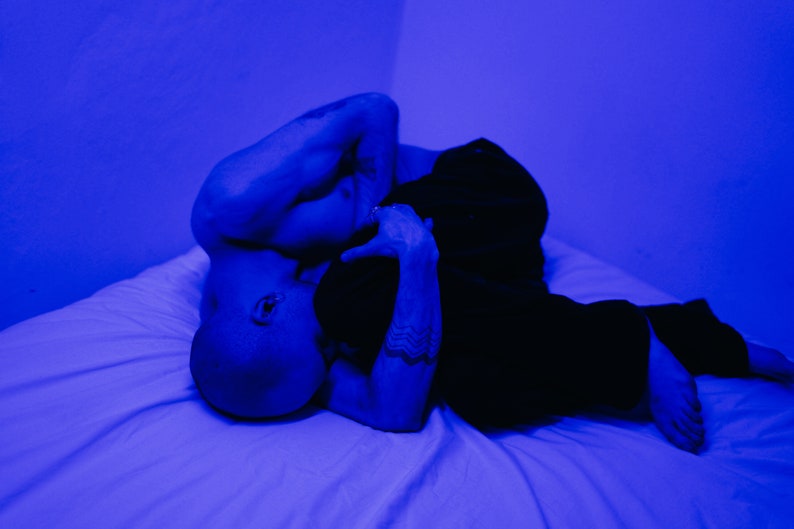May include: Two people lying in bed together, illuminated by blue light. One person is shirtless and the other is wearing black pants. The image has a moody and intimate feel.