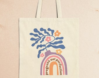 May include: A white canvas tote bag with a rainbow design and the text "progress not perfection" in a retro font. The rainbow is topped with a blue stylized plant with pink and orange flowers.