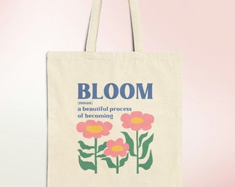 May include: A beige tote bag with a blue and white graphic that says "BLOOM (noun) a beautiful process of becoming" with three pink flowers with yellow centers and green leaves.