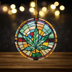 May include: Round Christmas ornament with a stained glass design featuring a green marijuana leaf in the center. The ornament is surrounded by a colorful circular pattern of red, yellow, blue, and green.
