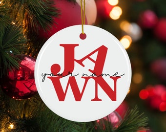 Op de afbeelding: Witte kerstbal van keramiek met de letters "JAwn" in rood en de tekst "your name" in cursief eronder.