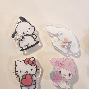 May include: Four clear acrylic pins featuring popular cartoon characters. Includes a white dog with black ears, a white bunny with a small dessert, Hello Kitty holding a red heart, and a pink character with a strawberry.