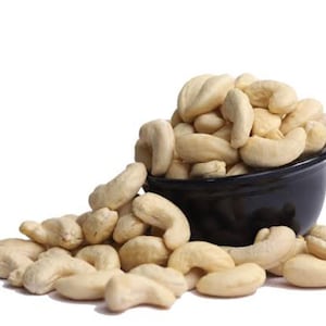 Puede incluir: Un cuenco negro rebosante de anacardos crudos, con anacardos adicionales esparcidos sobre una superficie blanca. Los anacardos son de color beige claro y tienen forma curva.