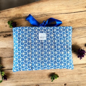 Può includere: Un oggetto in tessuto blu e bianco a motivi geometrici, forse una custodia o un astuccio, legato con un nastro blu. L'oggetto ha una piccola etichetta bianca con testo. Lo sfondo è una superficie di legno con piccoli fiori.