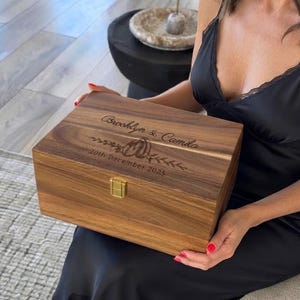 May include: A rectangular wooden keepsake box with a hinged lid and a gold-colored metal latch. The lid is engraved with the text 'Brooklyn & Camila', a graphic of two pumpkins, and the date '20th December 2025' in a dark, script-style font.