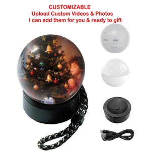 May include: A snow globe featuring a Christmas scene with a child, set on a black base with a black and white strap. Additional components include a white speaker, a charging base, and a USB cable. Text on the image reads: CUSTOMIZABLE, Upload Custom Videos & Photos, I can add them for you & ready to gift.
