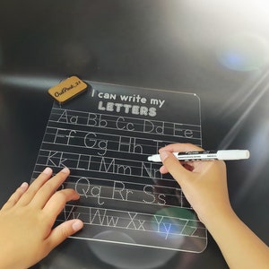 May include: A black dry-erase board with white lettering that says "I can write my LETTERS". The board has white dotted lines for tracing letters of the alphabet. A child's hand is holding a white dry-erase marker.