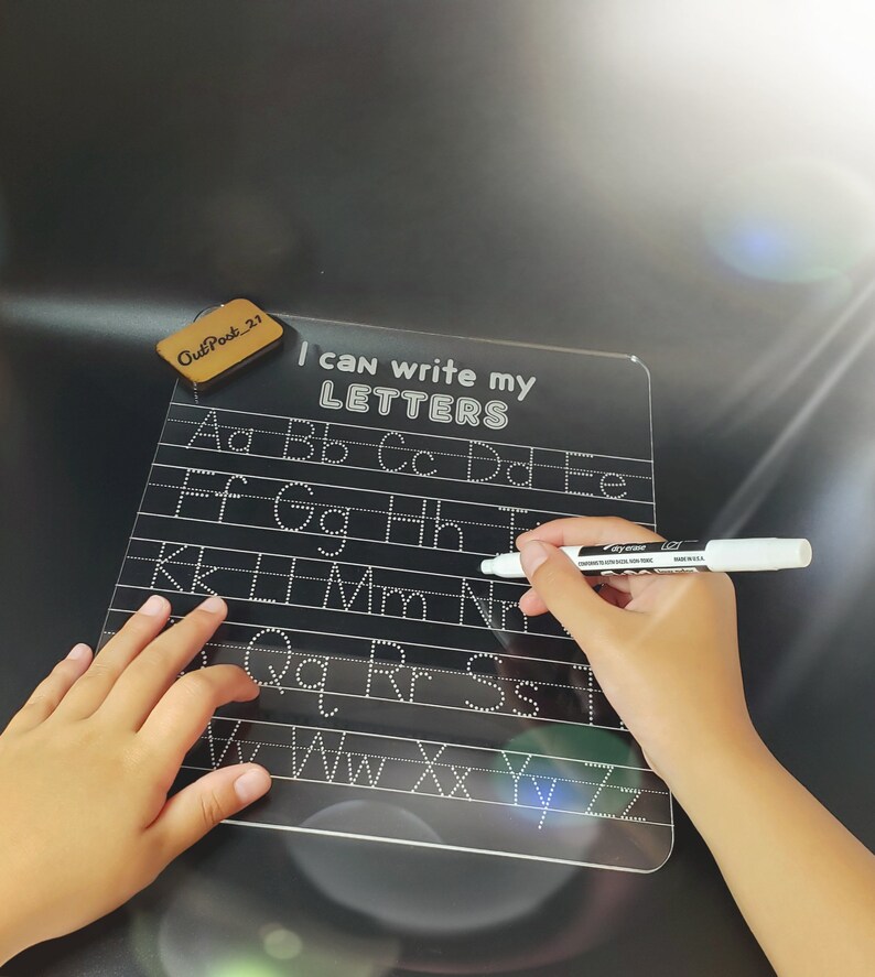 May include: A black dry-erase board with white lettering that says "I can write my LETTERS". The board has white dotted lines for tracing letters of the alphabet. A child's hand is holding a white dry-erase marker.