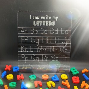 May include: A clear plastic dry-erase board with the text "I can write my LETTERS" at the top. The board has dotted lines for tracing the alphabet in uppercase letters.  A collection of colourful magnetic letters are scattered below the board.