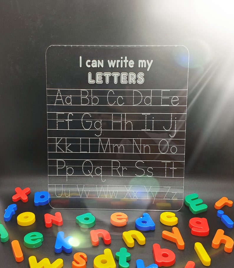 May include: A clear plastic dry-erase board with the text "I can write my LETTERS" at the top. The board has dotted lines for tracing the alphabet in uppercase letters.  A collection of colourful magnetic letters are scattered below the board.