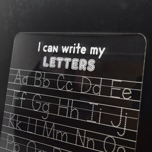 May include: A black dry-erase board with white dotted lines and letters for practicing writing the alphabet. The board has the text "I can write my LETTERS" at the top.