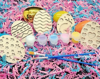 May include: A set of six wooden Easter egg shapes with different designs, including stars, hearts, waves, and a bunny. The eggs are ready to be painted with the included set of five paint pots and two paintbrushes. The eggs are surrounded by pink and blue crinkle paper.