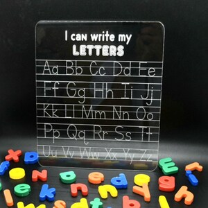 May include: A clear acrylic dry-erase board with the text "I can write my LETTERS" and dotted lines for tracing the alphabet.  The board is surrounded by colourful magnetic letters.