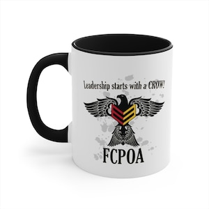 May include: White ceramic mug with a black handle and black rim. The mug features a black and white eagle with a red and yellow chevron on its chest. The text "Leadership starts with a CROW!" and "FCPOA" are printed on the mug.