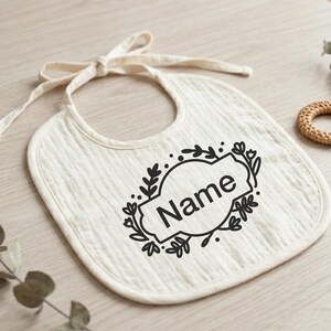 K&ouml;nnte beinhalten: Cremefarbenes Baby-L&auml;tzchen mit Bindeband. Das L&auml;tzchen hat ein schwarzes Blumendesign um ein Oval mit dem Wort "Name" in Schwarz gedruckt. Das L&auml;tzchen ist aus einem weichen, strukturierten Stoff.
