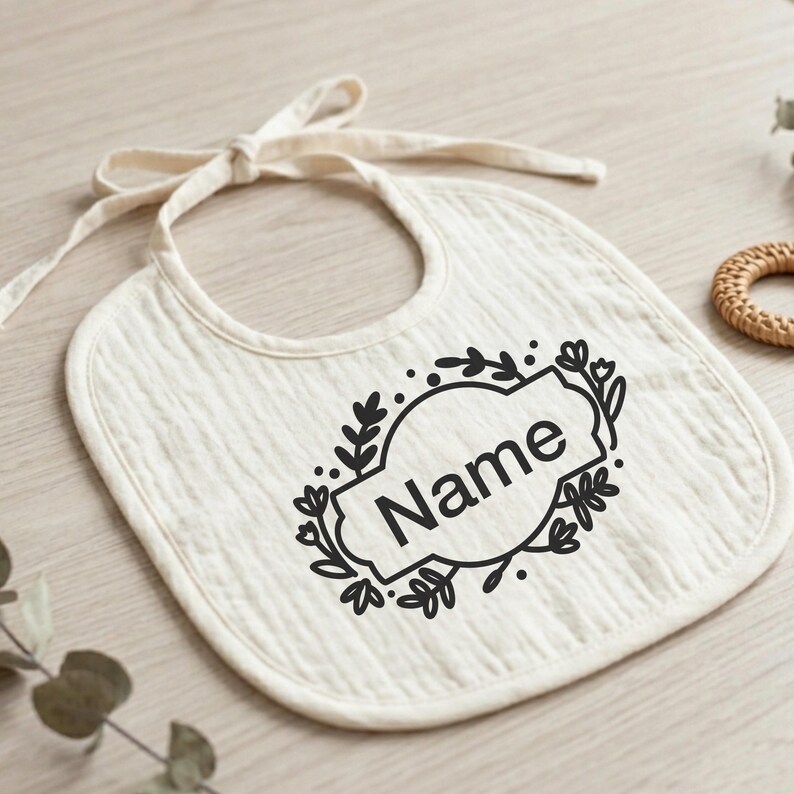 K&ouml;nnte beinhalten: Cremefarbenes Baby-L&auml;tzchen mit Bindeband. Das L&auml;tzchen hat ein schwarzes Blumendesign um ein Oval mit dem Wort "Name" in Schwarz gedruckt. Das L&auml;tzchen ist aus einem weichen, strukturierten Stoff.
