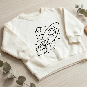 Könnte beinhalten: Cremefarbenes Sweatshirt mit einem schwarzen Raketendesign, einem Planeten und Sternen. Die Rakete hat ein kreisförmiges Fenster und Flammen. Das Sweatshirt hat einen Rundhalsausschnitt und gerippte Bündchen und Saum. Bequemes Kleidungsstück für Kinder.