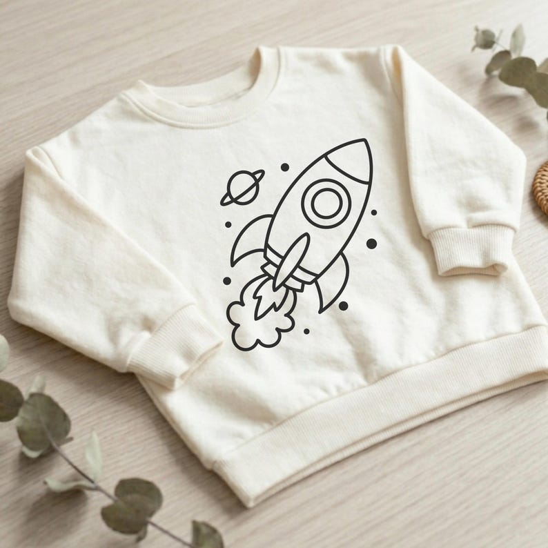 K&ouml;nnte beinhalten: Cremefarbenes Sweatshirt mit einem schwarzen Raketendesign, einem Planeten und Sternen. Die Rakete hat ein kreisf&ouml;rmiges Fenster und Flammen. Das Sweatshirt hat einen Rundhalsausschnitt und gerippte B&uuml;ndchen und Saum. Bequemes Kleidungsst&uuml;ck f&uuml;r Kinder.