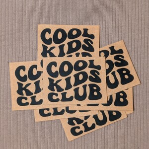 Könnte beinhalten: Mehrere braune, rechteckige Aufkleber mit dem Schriftzug "COOL KIDS CLUB" in schwarzer Retro-Schrift sind auf einer strukturierten, beigefarbenen Oberfläche angeordnet. Die Aufkleber überlappen sich leicht.