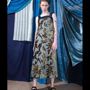 May include: A person wearing a long, blue and gold patterned dress with black straps. The dress has a floral design. The person is standing in front of a blue and white patterned background.