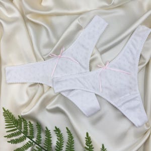 Puede incluir: Dos tangas blancas de encaje con lazos rosas. La ropa interior está colocada sobre una tela de satén blanco.