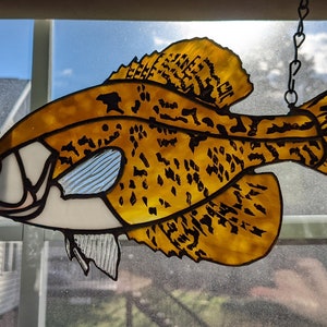 May include: Stained glass fish decoration in shades of yellow, white, and black, hanging from a chain. The fish has a detailed pattern and is displayed in front of a window.