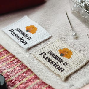 Könnte beinhalten: Zwei quadratische Stoffetiketten mit dem Text "HANDMADE BY Passion" und einem floralen Design. Ein Etikett ist weiß, das andere beige. Eine rote Garnrolle, Nadeln und Stoff sind ebenfalls sichtbar.