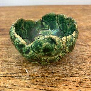 Puede incluir: Un pequeño cuenco de cerámica hecho a mano con un diseño de caparazón de tortuga. El cuenco es principalmente verde con detalles en verde oscuro y marrón. El borde tiene un borde texturizado e irregular. El cuenco está sobre una superficie de madera.