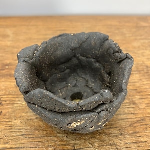May include: A small, handmade, black ceramic bowl with a rough, textured surface. The bowl has an irregular, organic shape with a dark interior and flecks of gold. The rim is uneven, adding to its rustic charm.