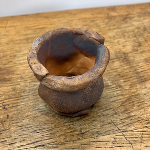 Può includere: Un piccolo vaso in ceramica marrone fatto a mano con una superficie strutturata. Il vaso ha una base arrotondata e un bordo ampio e irregolare. L'interno è di colore marrone chiaro. Il vaso viene probabilmente utilizzato per piantare piccole piante grasse o erbe aromatiche.