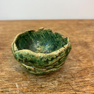Può includere: Una piccola ciotola in ceramica fatta a mano con un design strutturato e organico. La ciotola presenta una glassa verde screziata con sfumature di giallo e marrone, creando un'estetica naturale e terrosa. Il bordo ha una forma leggermente irregolare.