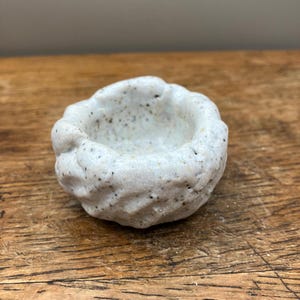 Könnte beinhalten: Eine kleine, weiße Keramikschale mit einer strukturierten, rauen Oberfläche. Die Schale hat eine leicht abgerundete Form und einen kleinen, flachen Innenraum.