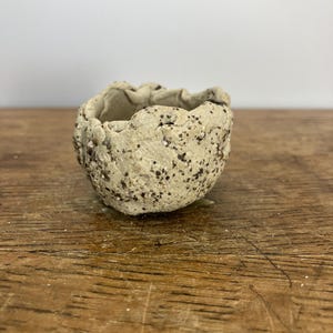 May include: A small, handmade, beige bowl with a rough, textured surface. The bowl has a rounded shape with an uneven, wavy rim. Dark speckles are scattered across the exterior. The bowl sits on a wooden surface.