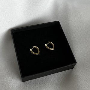 Könnte beinhalten: Ein Paar kleine, goldfarbene Creolen in einer schwarzen Schachtel. Die Ohrringe haben ein einzigartiges, eckiges Design. Die Schachtel befindet sich vor einem weißen, strukturierten Hintergrund.